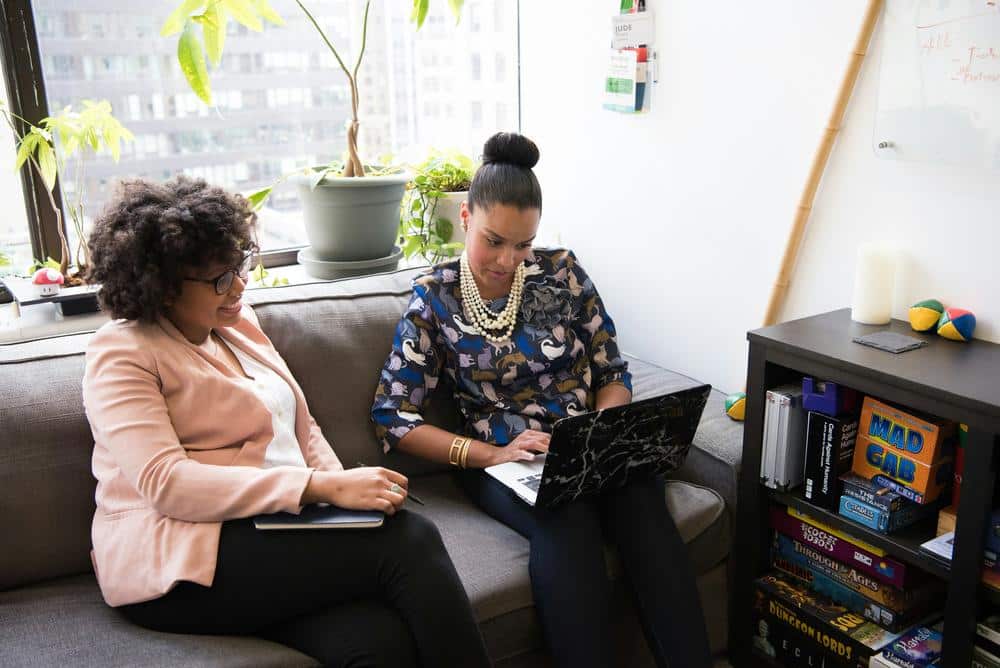 Mujeres en un sofá en un ambiente interior acogedor, con decoración moderna y plantas verdes alrededor.